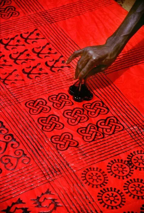 A hand lifting a carved stamp from red cloth printed in black with rows of interlaced knot motifs, wheel motifs and other Adinkra symbols.