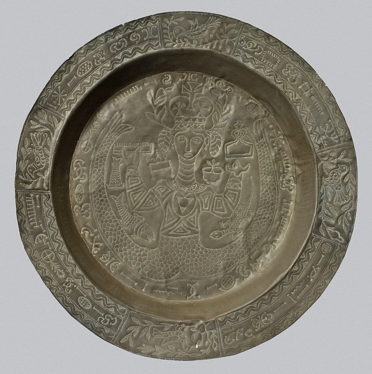 A round hammered brass dish: a crowned mermaid holding objects at the centre, surrounded by fish scales, birds and Nsibidi signs in panels around the rim.