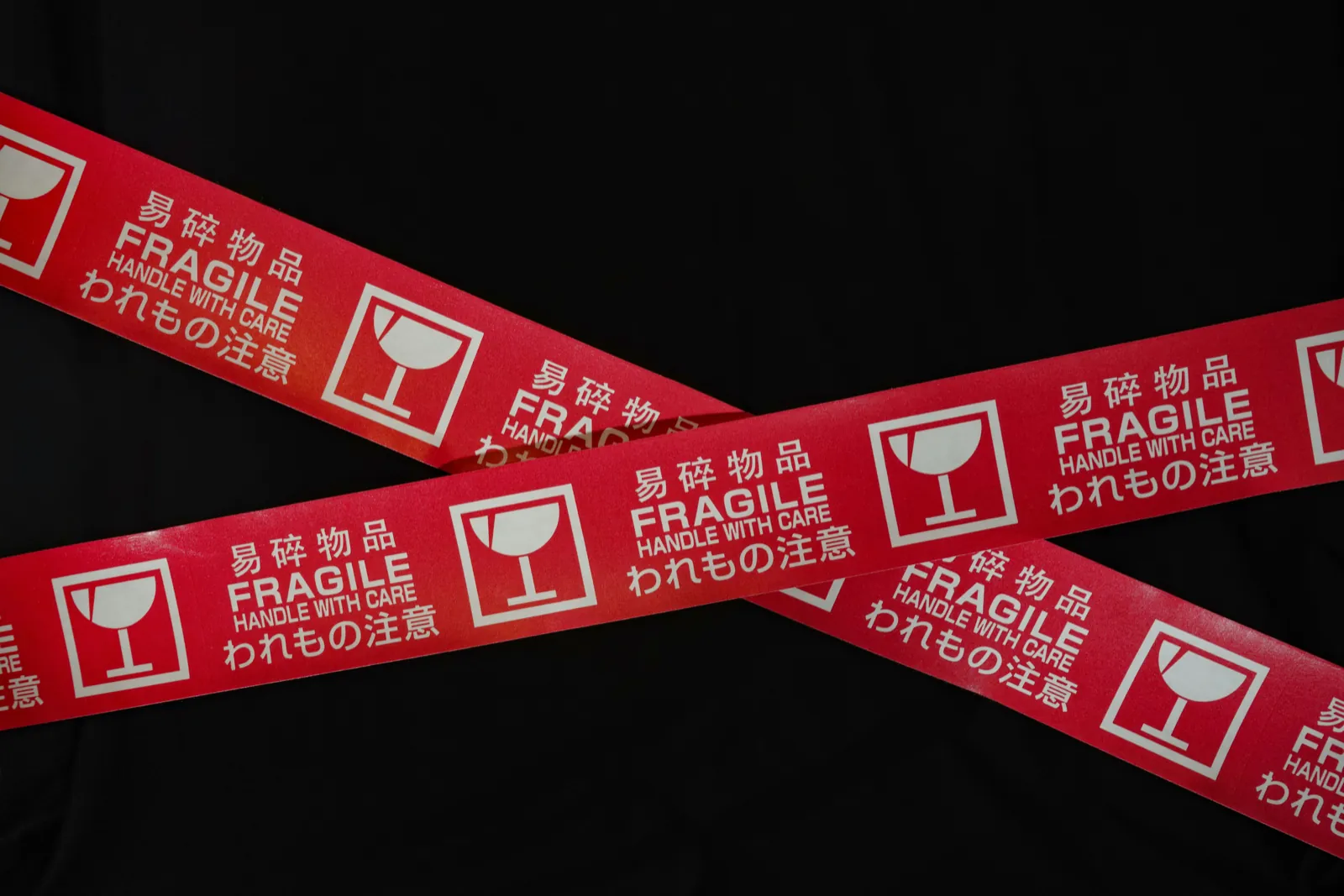 Two strips of red “Fragile, handle with care” tape crossing on black, printed with a broken-glass symbol and Chinese and Japanese text.